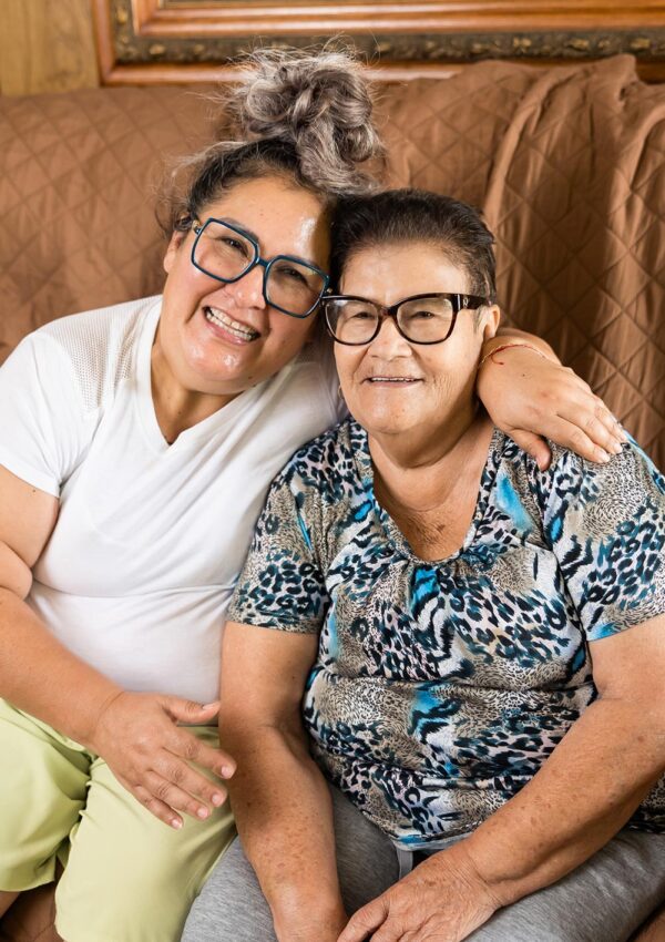 an elderly woman and her caregiver sitting on the couch and smiling together