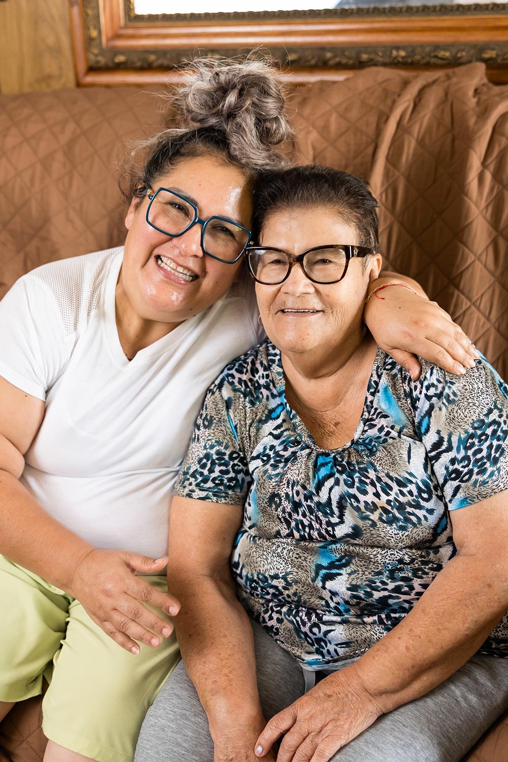 an elderly woman and her caregiver sitting on the couch and smiling together
